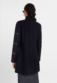 Abrigo de lana azul marino con tela de cuadros en las mangas, que presenta un cuello alto y una silueta entallada. Textura suave con detalles de costuras sutiles.