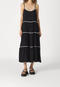 Robe maxi noire à volants avec un bord festonné blanc, fine bretelles et tissu léger. Associée à des sandales noires à plateforme épaisses.
