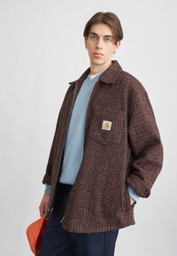 Brun og rød ternet uldjakke med lynlås fortil, oversized pasform, sidelommer og en brystlappe. Båret over en blå sweater og marineblå bukser.