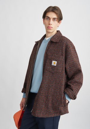 Brun og rød ternet uldjakke med lynlås fortil, oversized pasform, sidelommer og en brystlappe. Båret over en blå sweater og marineblå bukser.