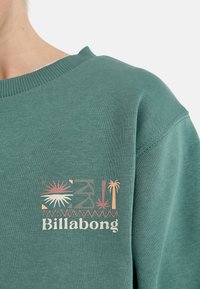 Sudadera de algodón verde con un logo impreso que presenta motivos de palmeras y sol en tonos rosa y beige en la parte izquierda del pecho. El texto dice "Billabong."