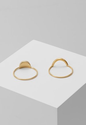 To tynde guldringe med hævede, afrundede geometriske former på en hvid pedestal mod en grå baggrund.