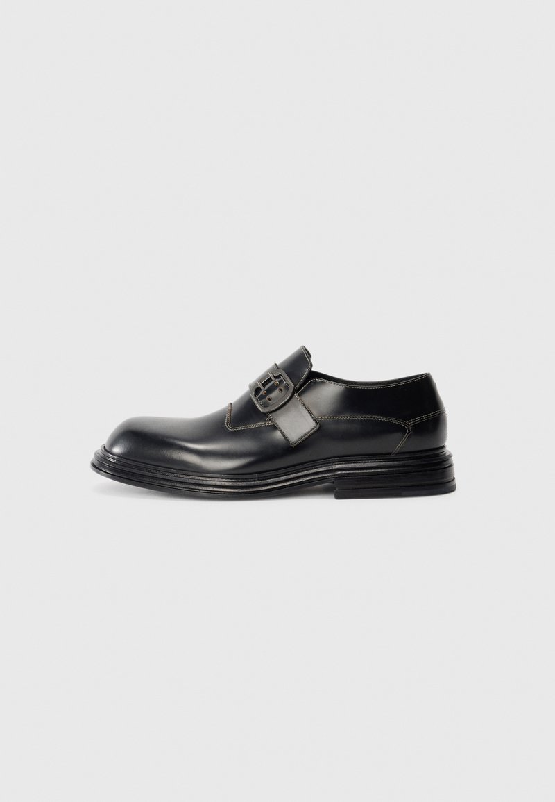 Chaussure habillée en cuir noir avec une large fermeture à boucle, des coutures détaillées et une semelle superposée, photographiée sur un fond blanc.