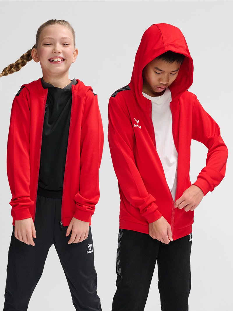Dwoje dzieci noszą czerwone bluzy na suwak i czarne spodnie sportowe; jedno się uśmiecha z opuszczonymi rękami, drugie zasuwa bluzę z podniesionym kapturem.