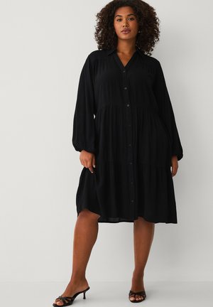 Kvinde med krøllet hår iført en sort midi-kjole med lange ærmer og knapper samt sorte hælede sandaler, stående mod en hvid baggrund.