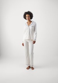 Vitt pyjamasset med svarta detaljer. Överdelen har långa ärmar, krage och en ficka. Byxorna är hel Längd och något utsvängda.