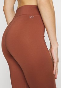 Leggings taille haute, ajustés, dans une couleur rouille. Fabriqués en tissu extensible, avec un petit logo blanc près de la taille. Texture lisse.