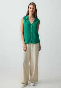 Donna in piedi che indossa una camicetta verde senza maniche, pantaloni beige a gamba larga e sneaker bianche su uno sfondo chiaro e semplice.
