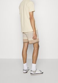 Hombre con pantalones cortos a rayas beige, camiseta crema, calcetines blancos y sandalias grises de Hugo, de pie frente a un fondo blanco liso.