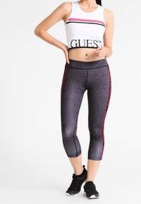 Vit croppad topp med ordet "GUESS" i svart, i kombination med svarta mönstrade capri leggings med röda sidostråk, bärs med svarta sneakers.