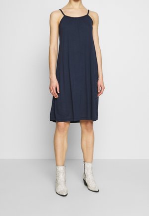 Kobieta ubrana w granatową, bezrękawną sukienkę do kolan z cienkimi ramiączkami oraz białe, wzorzyste kozaki sięgające połowy łydki, stojąca na tle jednolitego tła.