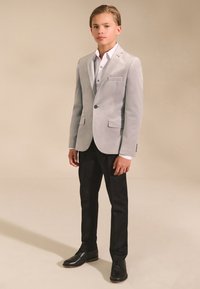 Hellgrauer Samtblazer mit zwei Vorderentaschen, kombiniert mit einem weißen Hemd und schwarzen Hosen. Schwarze Herrenschuhe vervollständigen das Outfit.