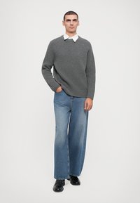 Pull tricot gris à texture côtelée, associé à un jean en denim bleu à coupe ample. Le modèle porte également une chemise blanche à col et des chaussures noires.