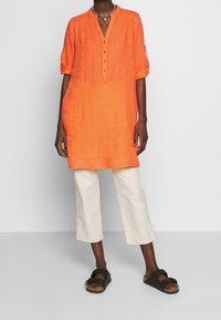 Tunica di lino arancione con scollo a V e tre bottoni, abbinata a pantaloni cropped crema e sandali neri. Design casual e comodo.