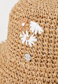 Geweven strohoed in tan met witte en bruine bloemenversieringen, voorzien van een zilveren logo hardware detail. Gelaagd en lichtgewicht ontwerp.