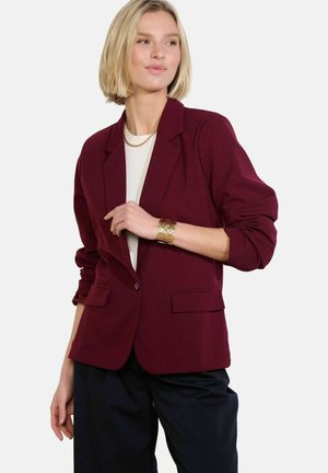 Donna con capelli biondi corti che indossa un blazer bordeaux, una maglietta bianca, pantaloni neri, una collana a catena d'oro e un braccialetto d'oro su uno sfondo semplice.