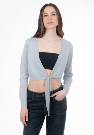 Femme aux longs cheveux bruns portant un cardigan court gris clair noué à la taille par-dessus un haut bandeau noir et un jean noir, avec une main dans la poche.