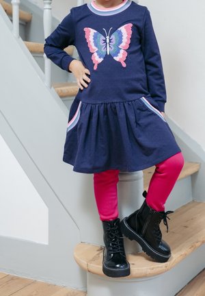 Marineblauwe jurk met een vlinderdesign, ronde hals, lange mouwen en een pofrok; gecombineerd met roze legging en zwarte veterlaarzen tot aan de enkel.