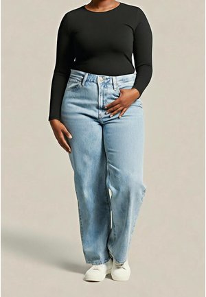 Vrouw draagt een zwarte top met lange mouwen, lichtblauwe high-waisted wijde jeans en witte sneakers, staand tegen een neutrale achtergrond.