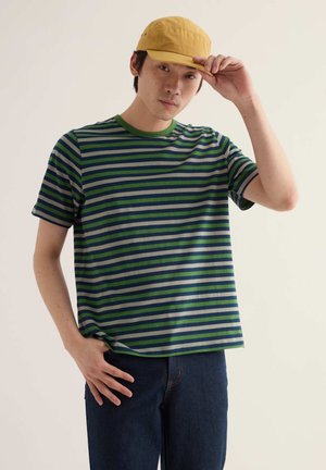 Mann mit grün-blau gestreiftem T-Shirt, dunklen Jeans und gelber Mütze, der mit einer Hand den Schirm der Mütze hält.