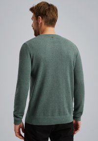 Pull en tricot vert, à manches longues, col rond, avec un petit logo dans le dos. Détails brodés aux poignets et à l'ourlet.