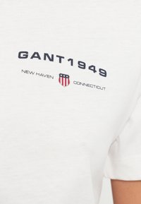 Bela bombažna majica s temnomodrim tiskom "GANT1949" in "NEW HAVEN CONNECTICUT" ter ščitnim motivom v rdeči in modri barvi. Preprost okrogel izrez.