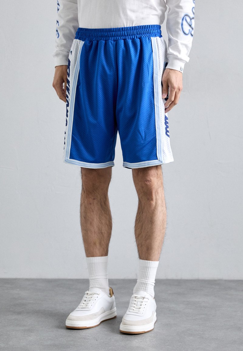 Blå mesh idrettsshorts med elastisk midje, hvite sidepaneler og logo på siden. Brukt med hvite joggesko og stripete sokker.