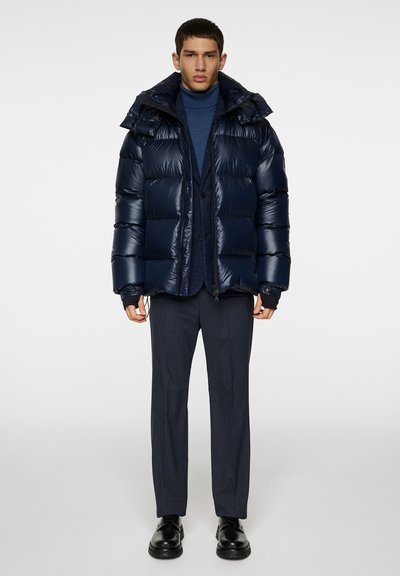 Navy puffer jacket with shiny, quilted texture, adjustable drawstring hood, and a front zipper. Worn over a blue turtleneck and grey trousers.
