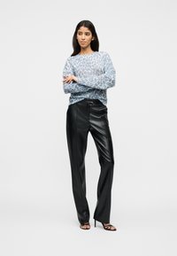 Γυναίκα όρθια με σταυρωμένα τα χέρια, φοράει ένα ανοιχτό μπλε πουλόβερ με λεοπάρ σχέδιο, μαύρο δερμάτινο παντελόνι και μαύρες ψηλοτάκουνα σανδάλια.