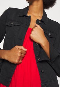 Veste en jean noire avec col classique et fermeture à boutons, portée par-dessus une robe rouge. Dotée de poches et d'une coupe cintrée. Tissu texturé.