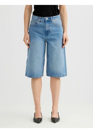 Mujer con pantalones cortos de mezclilla azul medio hasta la rodilla con bolsillos delanteros, combinados con tacones negros de punta afilada y una camiseta negra.