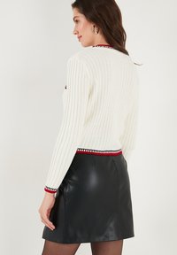 Bijela rebrasta jumper s crvenim i crnim prugastim detaljima na izrezima i manžetama, u kombinaciji s crnom kožnom mini suknjom. Teksturirana tkanina.