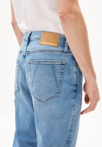 Jean bleu clair avec un design classique, style à cinq poches, et une patch en cuir marron avec "ARMEDANGELS" embouti au dos.