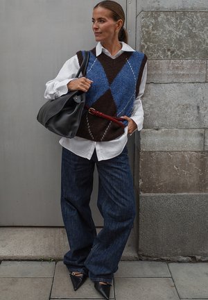 Bruine en blauwe argyle gebreide vest over een wit shirt, gecombineerd met wijde donkerblauwe denim jeans en zwarte puntige schoenen. Houdt een zwarte tas vast.