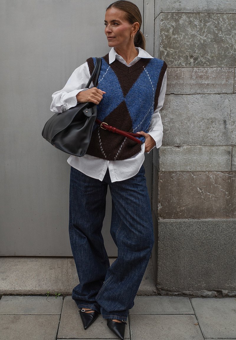 Chaleco de punto a rombos en marrón y azul sobre una camisa blanca, combinado con unos jeans de mezclilla oscuros de pierna ancha y zapatos negros de punta. Lleva un bolso negro.