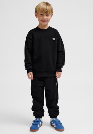 Sort sweatshirt og bukser sæt i blødt bomuldsstof med ribbede manchetter. Bemærkelsesværdig hvid logo detalje på brystet, parret med blå velcro sneakers.