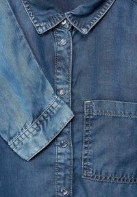 Modrá džínová košile s límcem na knoflíčky, dlouhými rukávy, přední kapsou a různorodými vzory blednutí na látce. Průhledné plastové knoflíky.