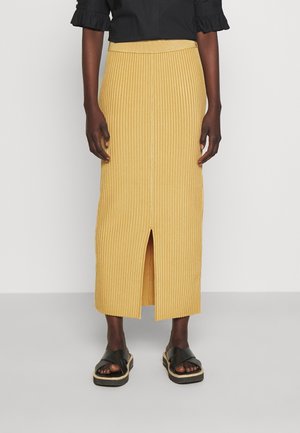 Żółta plisowana spódnica midi z przednim rozcięciem, z dopasowanym stanem. W połączeniu z czarnymi płaskimi sandałami. Tło jest jednolicie białe.