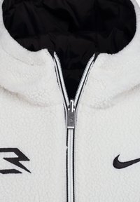 Veste zippée en polaire blanche avec doublure noire. Dispose d'une fermeture éclair avec des éléments en argent et des détails de logo noirs. Surface texturée.