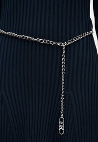 Αλυσίδα ασημένια ζώνη με λεπτομέρεια κούμπωμα λουκέτου που κρέμεται, φορεμένη πάνω από ένα σκούρο μπλε ριμπ πλεκτό ένδυμα.