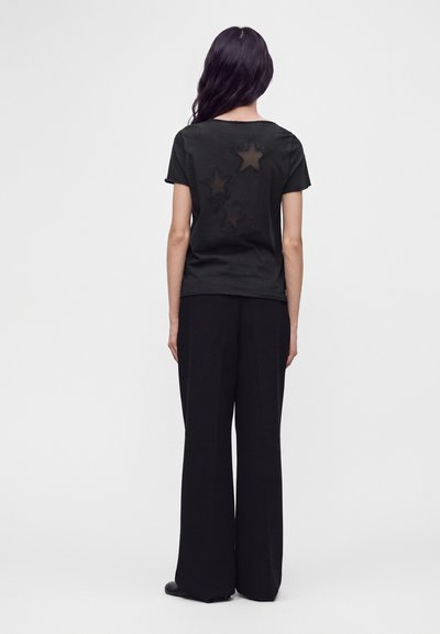 Woman with long dark hair stands facing away wearing black wide-leg pants and a black short-sleeve shirt with three star-shaped cutouts on the back.