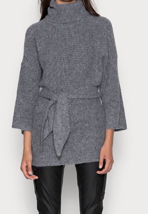 Femme portant un pull à col roulé gris en maille avec de larges manches et une ceinture assortie, associé à un pantalon en cuir noir.