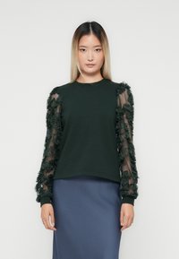 Donna con capelli ombre lisci che indossa un top verde scuro con maniche trasparenti a fantasia floreale e una gonna blu, su uno sfondo bianco semplice.