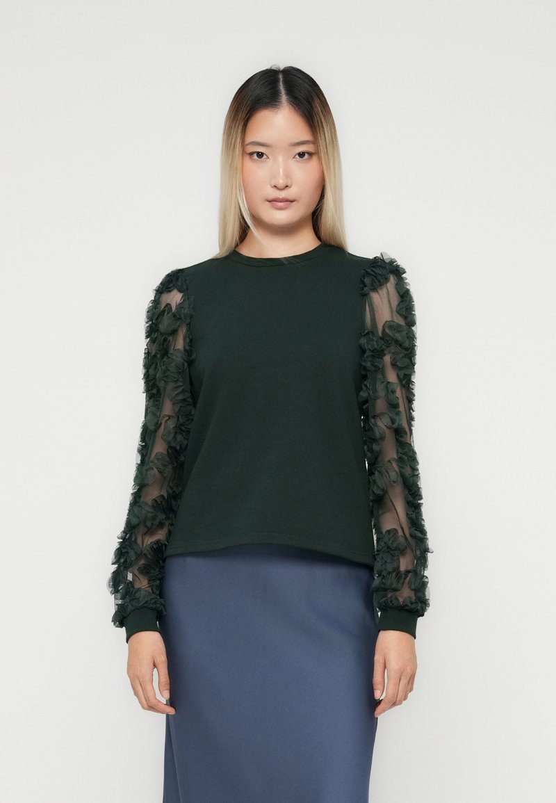 Donna con capelli ombre lisci che indossa un top verde scuro con maniche trasparenti a fantasia floreale e una gonna blu, su uno sfondo bianco semplice.