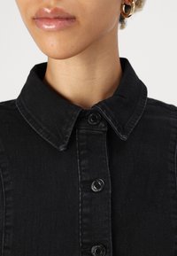 Crna traper košulja s ovratnikom, prednjim dugmadi i vidljivim šavovima. Glatka tekstura s strukturiranim dizajnom.