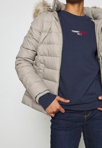 Giacca imbottita beige con cappuccio foderato in pelliccia, chiusura con zip e texture trapuntata, indossata sopra una felpa navy e jeans blu scuro.