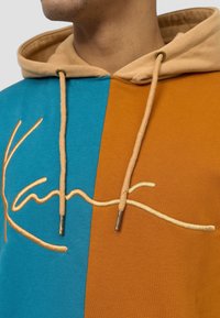 Felpa con cappuccio in tessuto blu e arancione, caratterizzata da un evidente logo oro in corsivo e coulisse beige con dettagli in metallo.