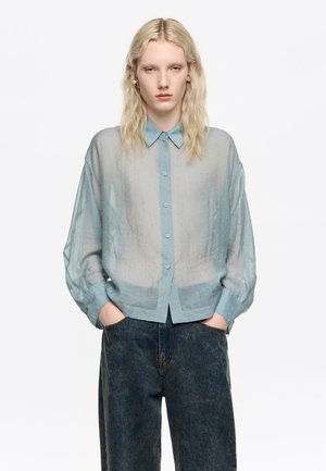 Camisa azul claro, semi-translúcida, con botones y cuello, con un patrón de puntos texturizados. Corte holgado con mangas abullonadas; combinada con unos jeans de mezclilla oscuros.