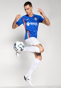 Modrá futbalová dres so červenými a žltými akcentmi, biele šortky a biele ponožky. Hráč udržuje vyšívanú futbalovú loptu v pohybe.