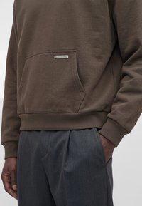 Personne portant un sweat-shirt marron avec une poche avant étiquetée "bazix republiq" et un pantalon plissé gris foncé, main dans la poche.